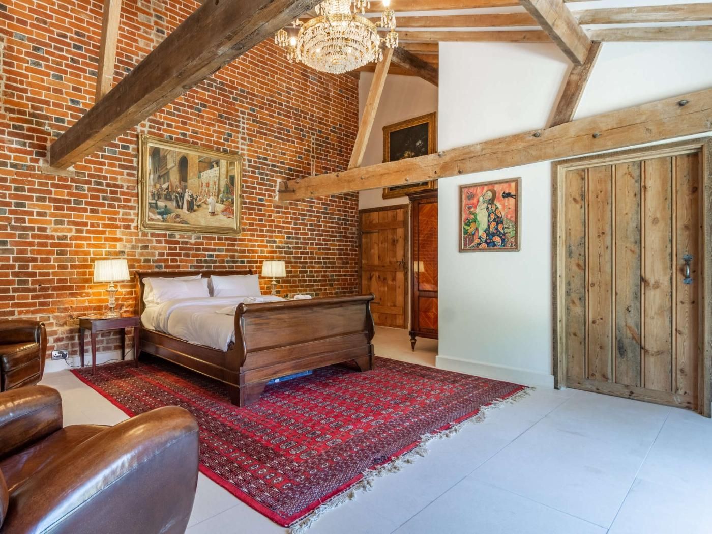 A bedroom with a brick wall, wooden ceiling beams, a bed, a red patterned rug, a wooden door, and a painting.