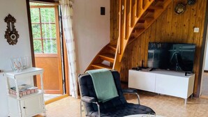 Ein Zimmer mit einer Holztreppe, einem Fernseher auf einem weißen Schrank, einem schwarzen Stuhl mit grüner Decke und einem weißen Tisch mit Gläsern.