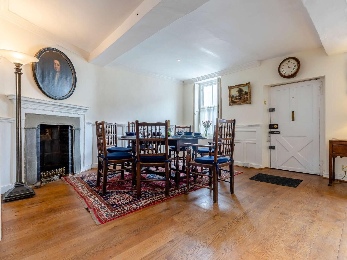 A dining room with a fireplace, wooden chairs, a table set for four, and a large painting on the wall.