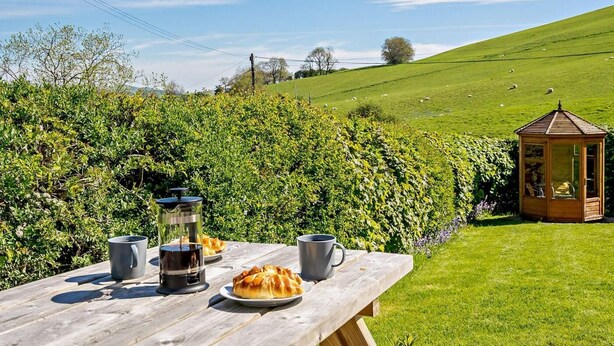 Une table de pique-nique avec de la nourriture et des boissons, un kiosque en bois et une colline verdoyante avec des moutons en arrière-plan.