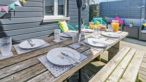 An outdoor dining area with a wooden table set for two, surrounded by a grey house with a window and a black pole. - 4 Bed in Newquay (oc-82951) (Newquay)