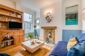 A cozy living room with a wooden bookshelf, a flat-screen TV, a coffee table with decorative items, a fireplace with a mirror above it, and a blue sofa. - 4 Bed in Newquay (oc-82951) (Newquay)