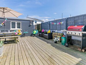 A wooden deck with a barbecue grill, picnic table, and outdoor seating area. - 4 Bed in Newquay (oc-82951) (Newquay)