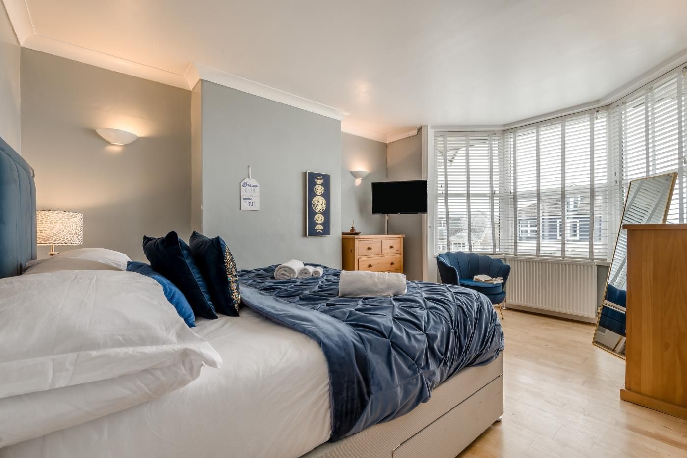 A bedroom with a bed, bedside table, dresser, TV, and a window with blinds.
