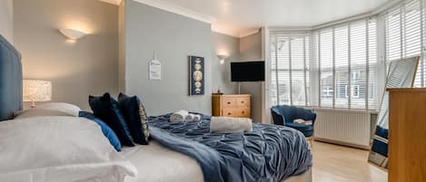 A bedroom with a bed, bedside table, dresser, TV, and a window with blinds.