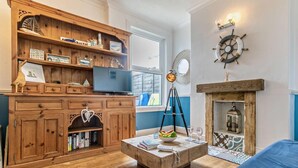 A cozy living room with a wooden entertainment unit, a television, a tripod lamp, a fireplace, and a blue sofa. - 4 Bed in Newquay (oc-82951) (Newquay)