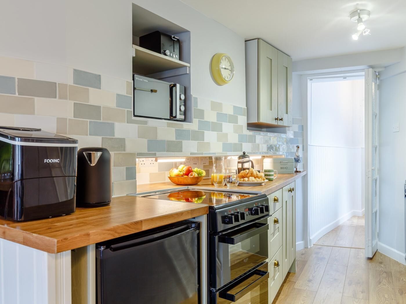 Una cocina moderna con una cafetera FoQING, un tostador, un lavabo y una estufa. Hay un reloj en la pared y una puerta que da a otra habitación.