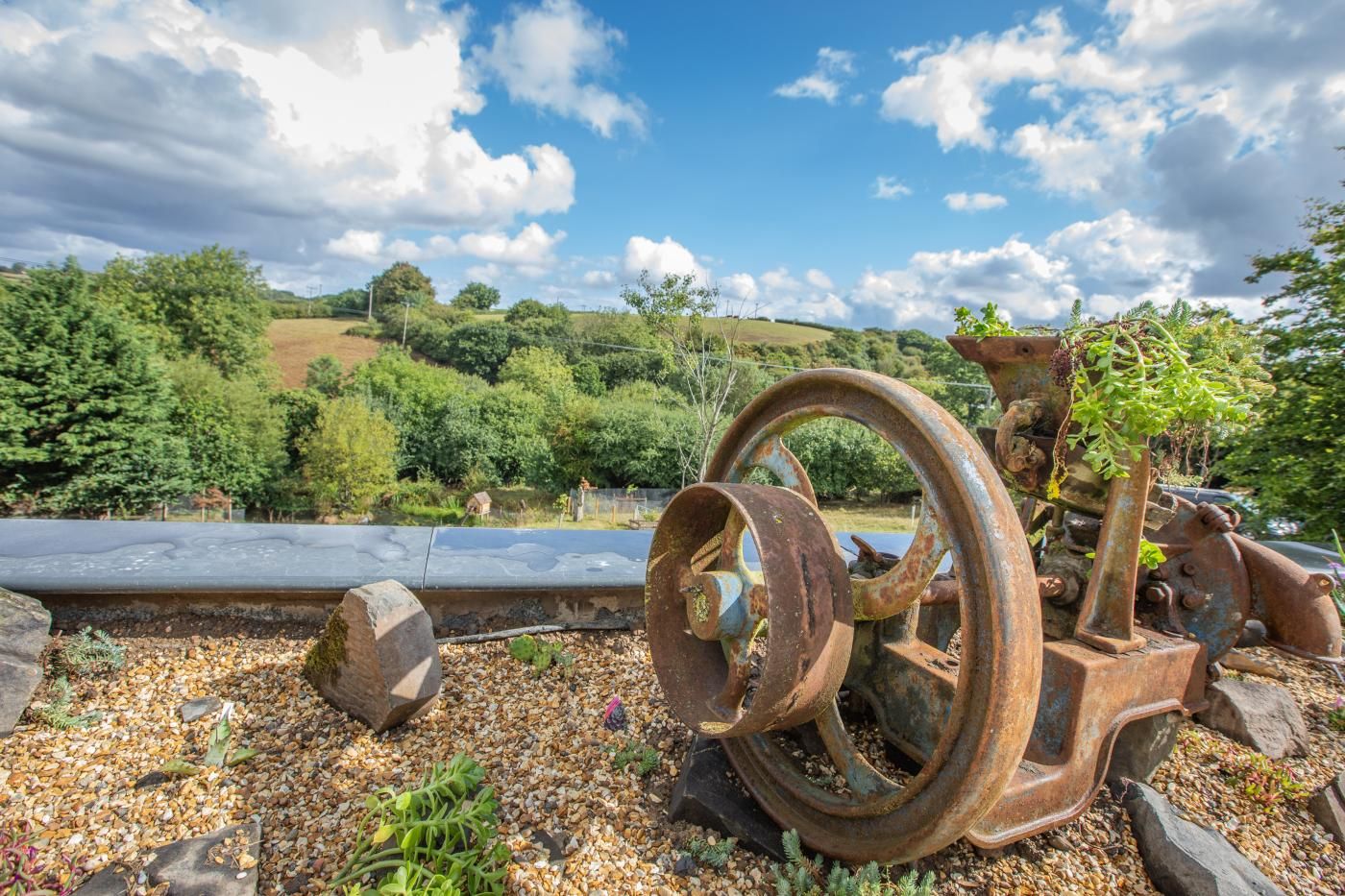An outdoor scene with a rusty piece of machinery, a gravel path, and a hilly landscape in the background.