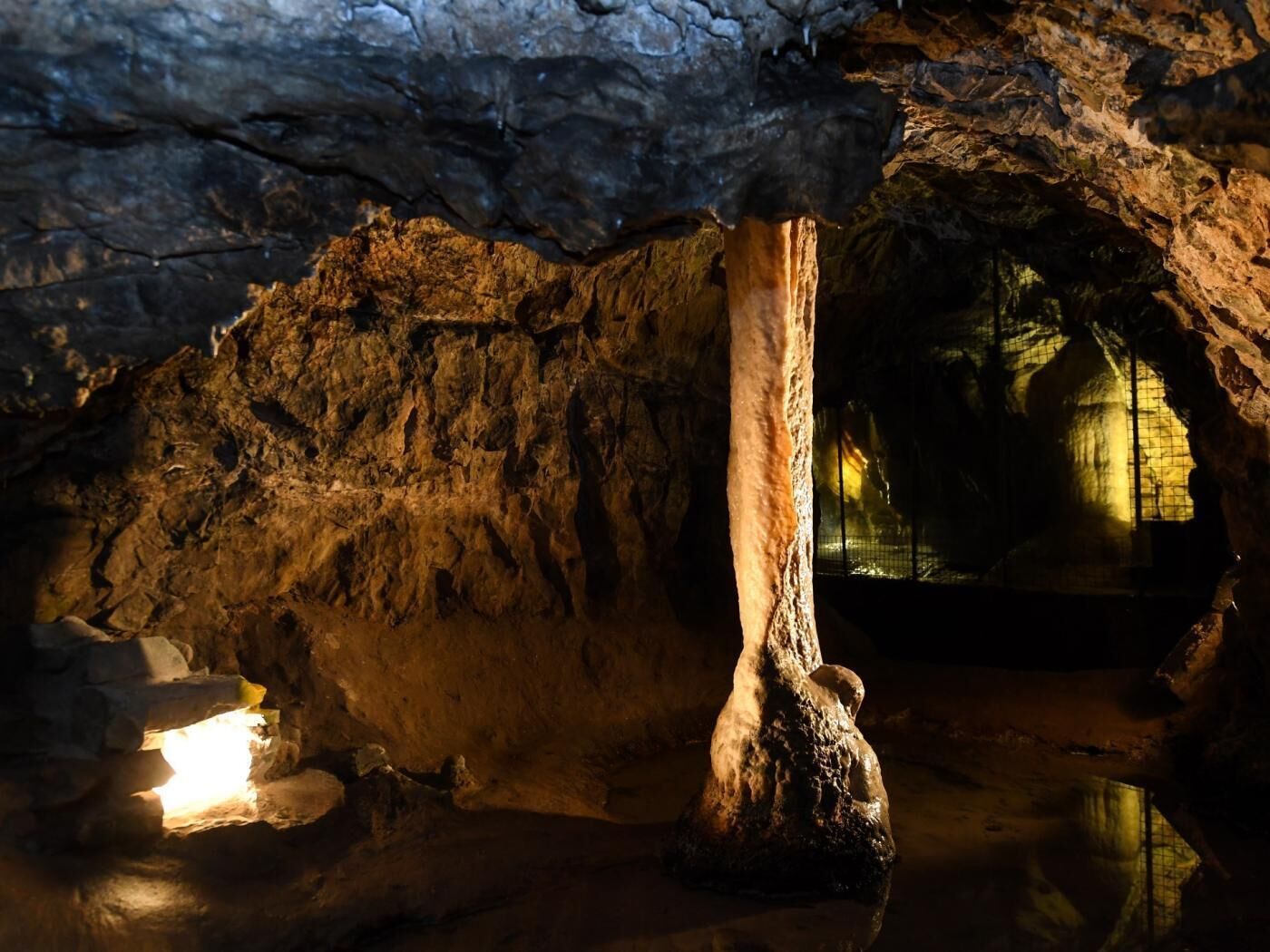 Una cueva con estalactitas, un pilar central y una zona iluminada al fondo.