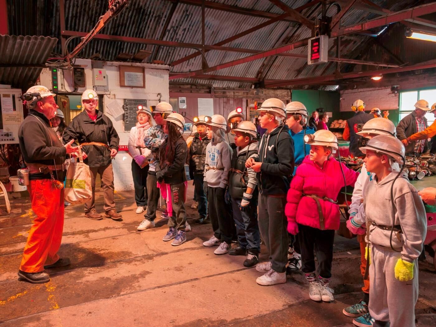 A group of people in hard hats and safety gear, standing in a line inside a building with a high ceiling and industrial features.