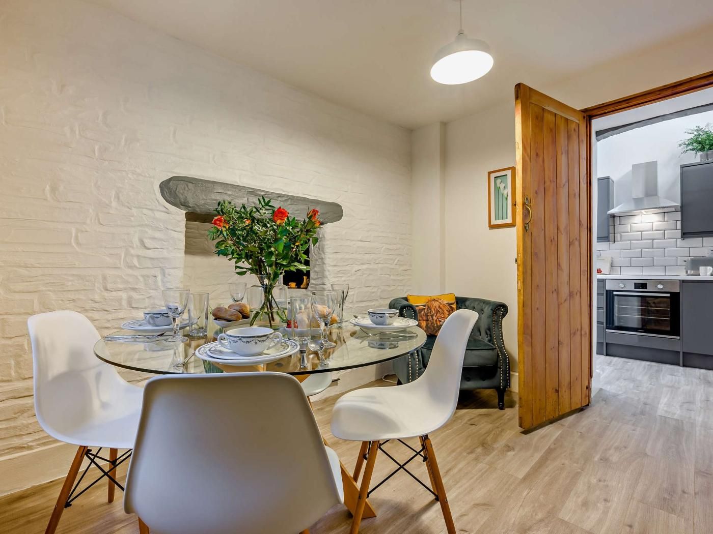 A dining area with a round glass table, white chairs, and a vase of flowers. There is a kitchen visible through an open door.