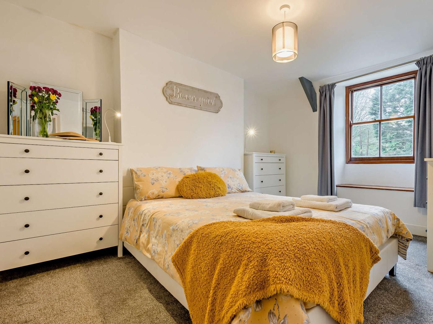 Une chambre à coucher avec un lit, une commode, une table de chevet, un miroir, une fenêtre avec des rideaux et un tableau accroché au mur.