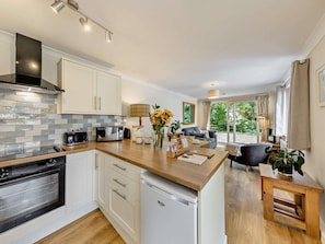 A modern kitchen with a dishwasher, a built-in oven, and a microwave. There is a dining area with a table and chairs, and a sofa. The room has a large window with curtains. - 2 Bed in Crewe (oc-83192) (Audlem)