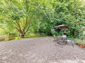 A gravel patio with a table and chairs, surrounded by dense foliage and a wooden fence. - 2 Bed in Crewe (oc-83192) (Audlem)