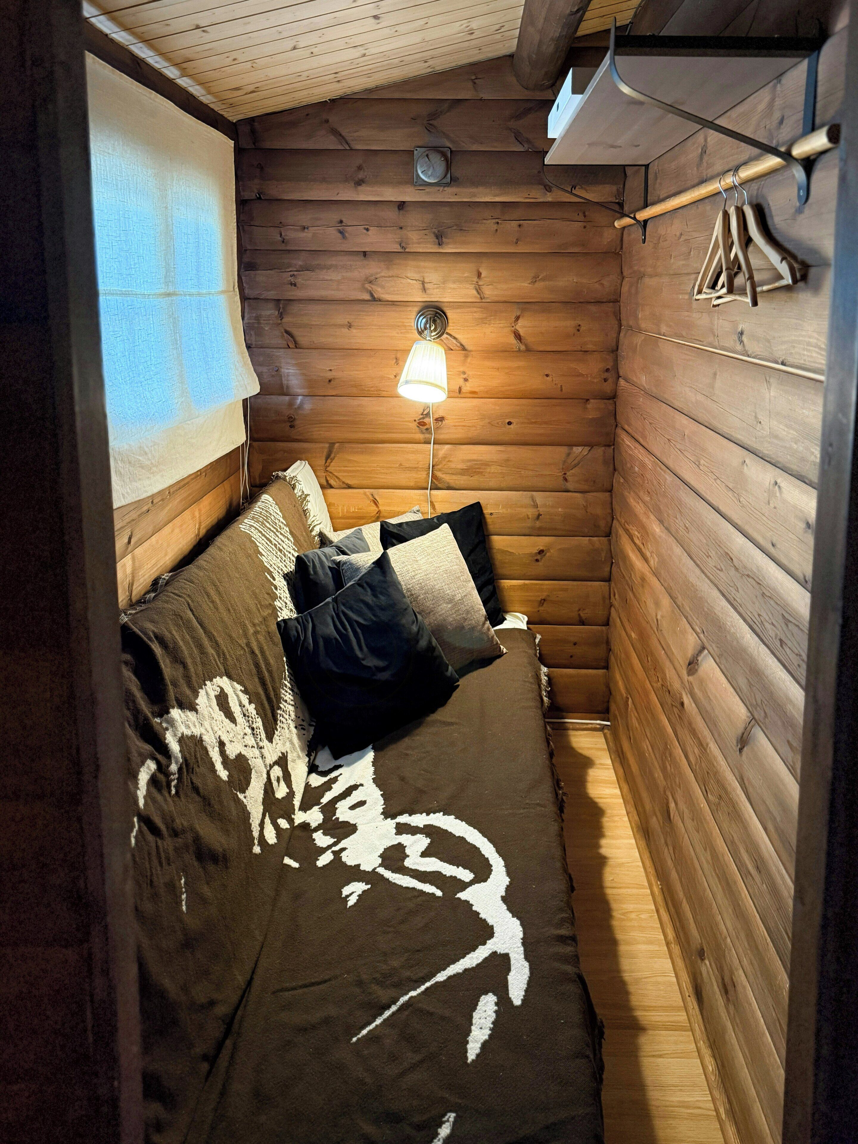 Um quarto pequeno e acolhedor, com decoração em madeira, uma cama com colcha estampada, uma luminária na parede e uma janela com cortina.