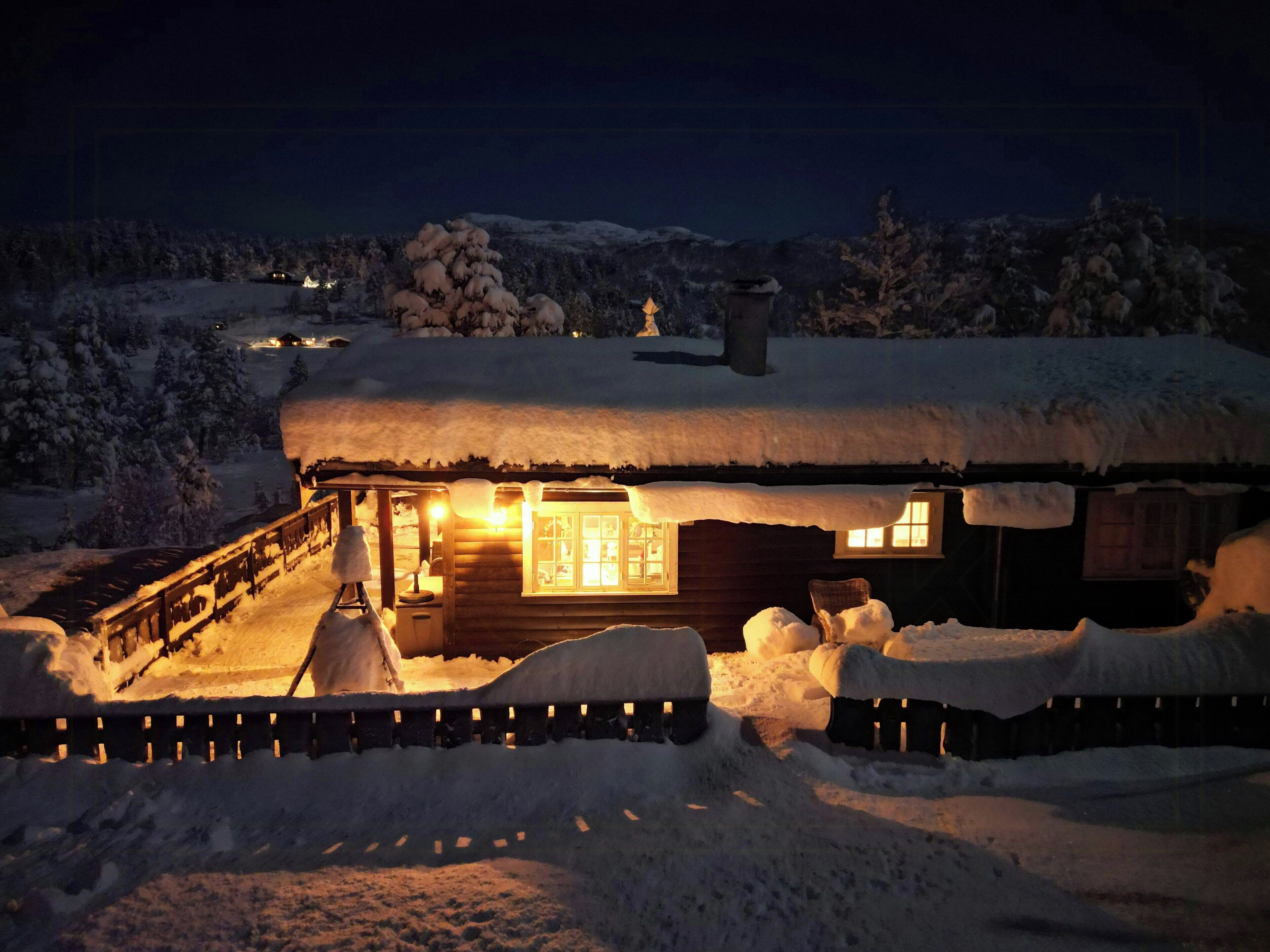 Une cabane en bois recouverte de neige, dont l’intérieur est éclairé, et qui est entourée de neige.