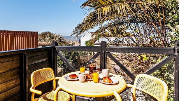 A wooden deck with a round table and two chairs, overlooking a beach and palm trees. - 2 Bed in Whitstable (oc-83291) (Whitstable)