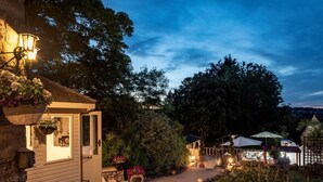 Ein Steinpfad führt zu einem Holzhäuschen mit einem hängenden Blumenkorb, einer Steinbank und einem kleinen Garten mit Kübelpflanzen und einer Pergola.