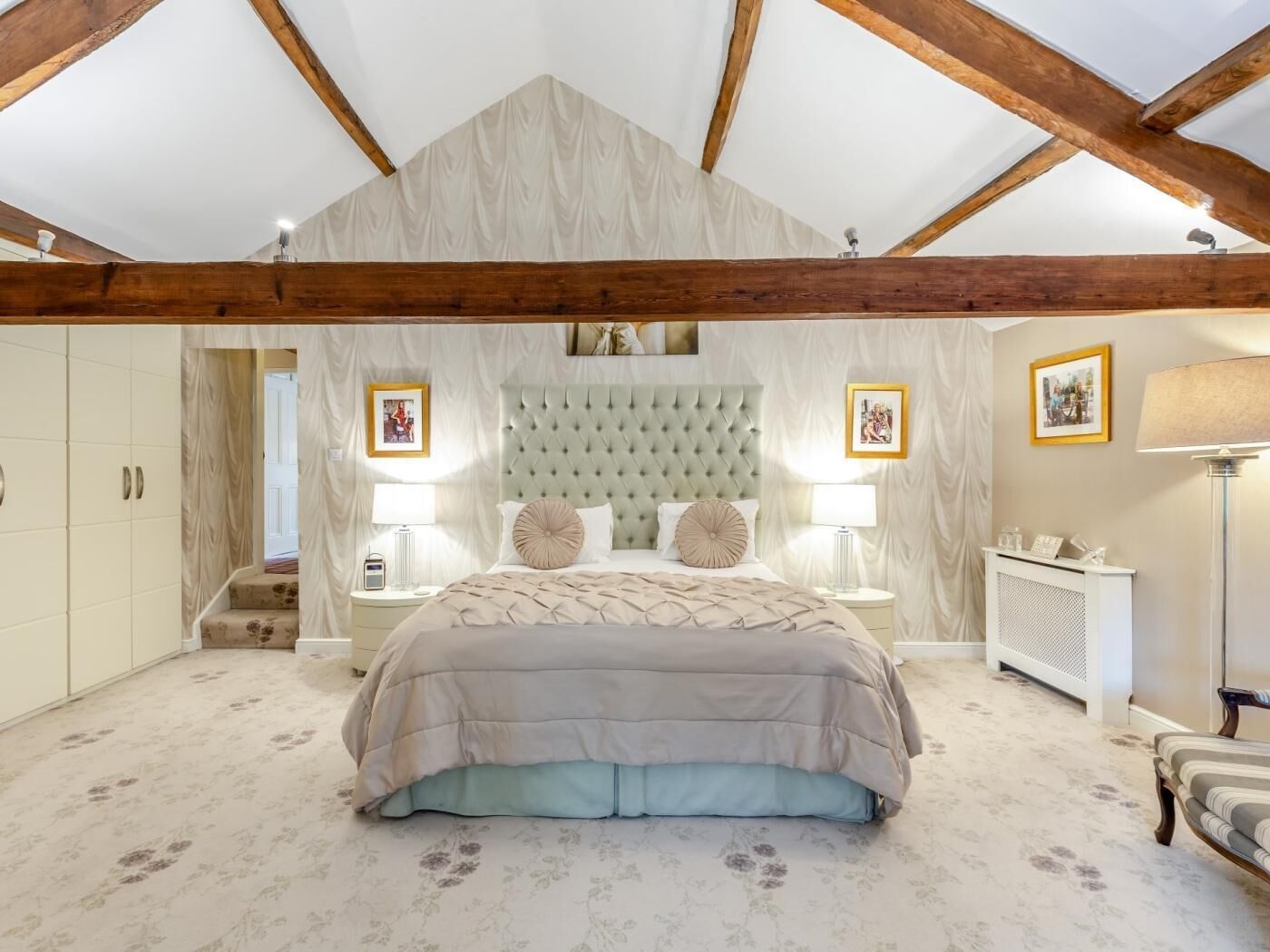 A bedroom with a large bed, wooden beams, a tufted headboard, bedside lamps, a nightstand, a radiator, and framed pictures on the wall.