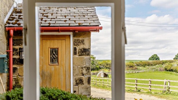 A stone cottage with a wooden door, a window with a diamond pattern, and a view of a rural landscape with a fence and greenery. - 3 Bed in Great Ayton (oc-83328) (Great Ayton)