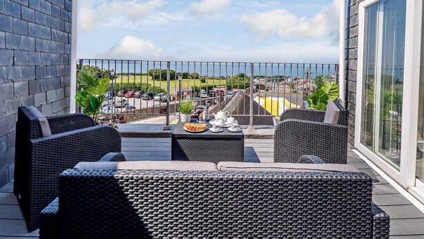 A balcony with wicker furniture, a grill, and a view of a parking lot and greenery. - 2 Bed in Woolacombe (oc-83425) (Woolacombe)