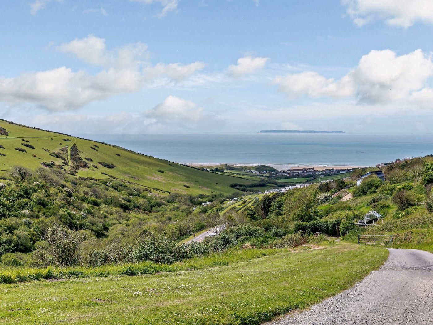 A rural landscape with a gravel road, green fields, and a distant view of the sea.