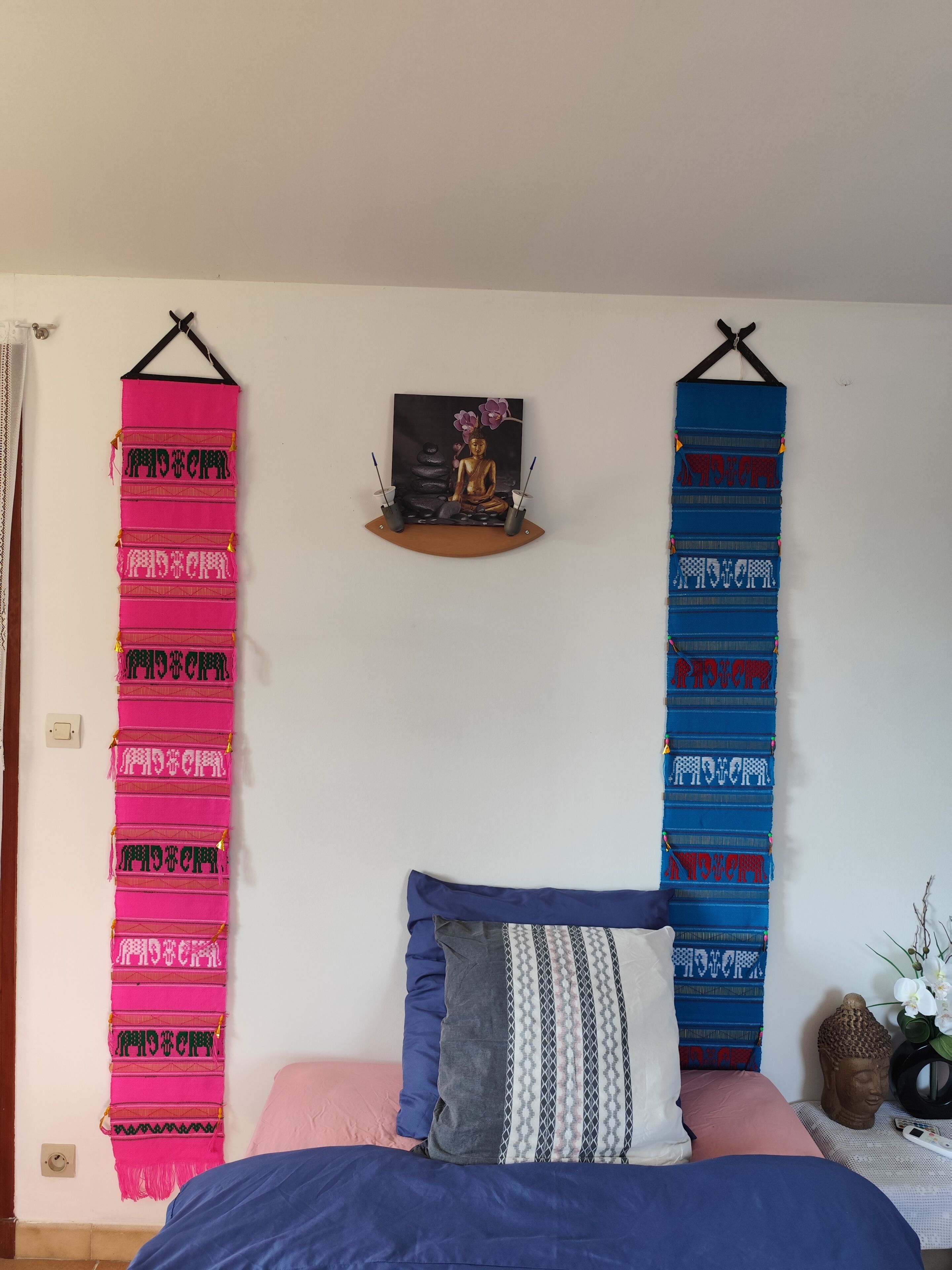 A bedroom with a bed, pillows, a wall-mounted shelf with a Buddha statue, and two vertical wall hangings.