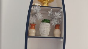 Hyllyllä on Buddhan patsas, kultainen esine ja koristeellisia kukkia.
