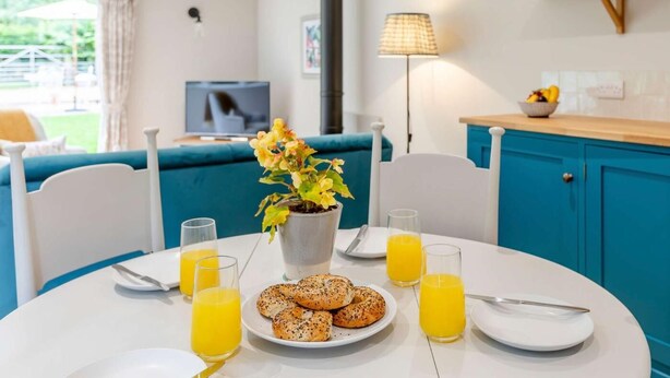 A dining area with a table set for breakfast, including bagels and orange juice, a flower vase, and a TV in the background. - 1 Bed in Wincanton (oc-83499) (Holton)
