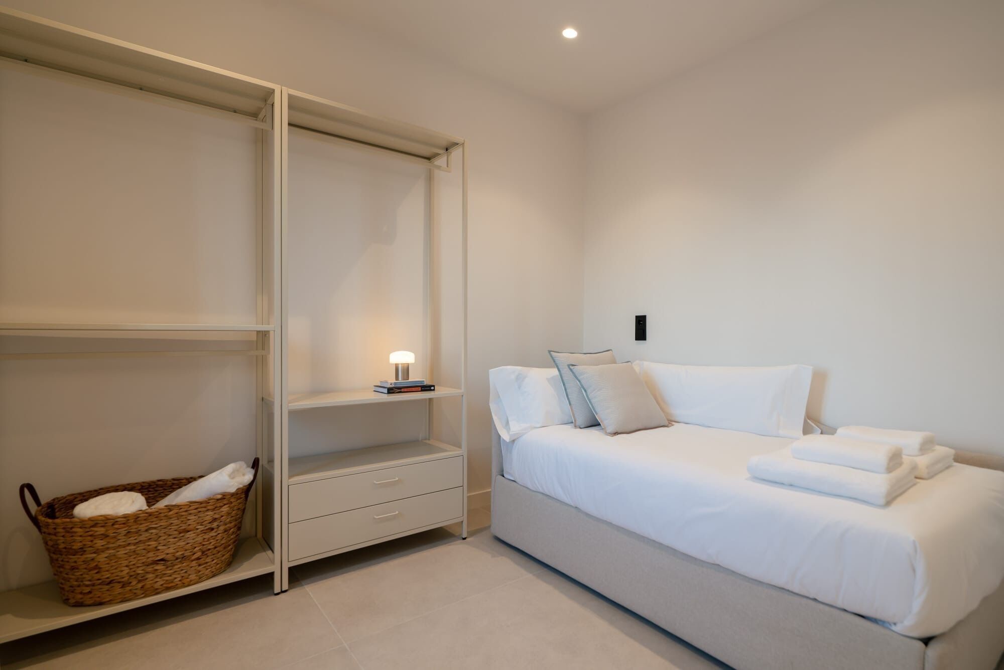 Een slaapkamer met een bed, nachtkastje, lamp, kast en een rieten mand.