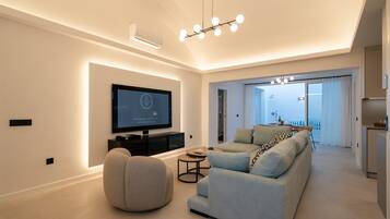 A modern living room with a flat-screen TV, a sofa, a round ottoman, and a dining area with a table and chairs.