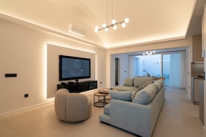 Ein modernes Wohnzimmer mit Flachbildfernseher, einer Couch, einem runden Hocker und einem Essbereich mit Tisch und Stühlen.