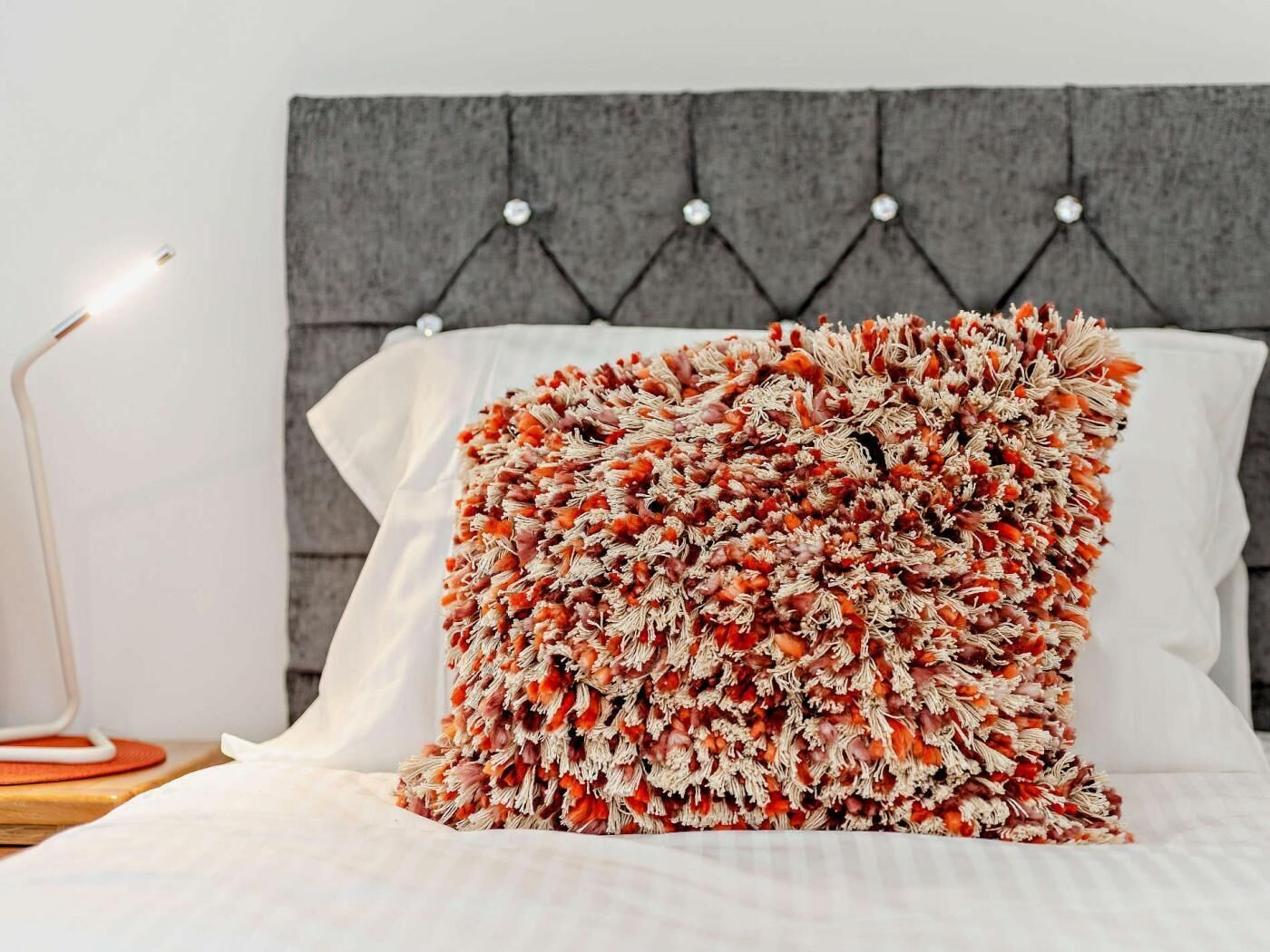 Una cama con un cojín decorativo, una lámpara de noche y un cabecero tapizado en gris.