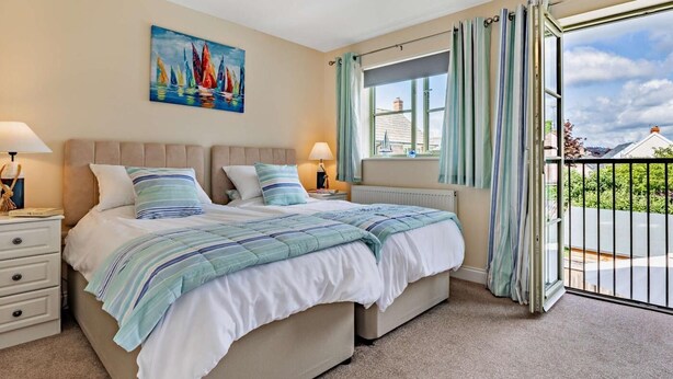 A bedroom with a large bed, two bedside tables with lamps, a painting on the wall, a window with curtains, and a view of the outside. - 3 Bed in Watchet (oc-83532) (Watchet)