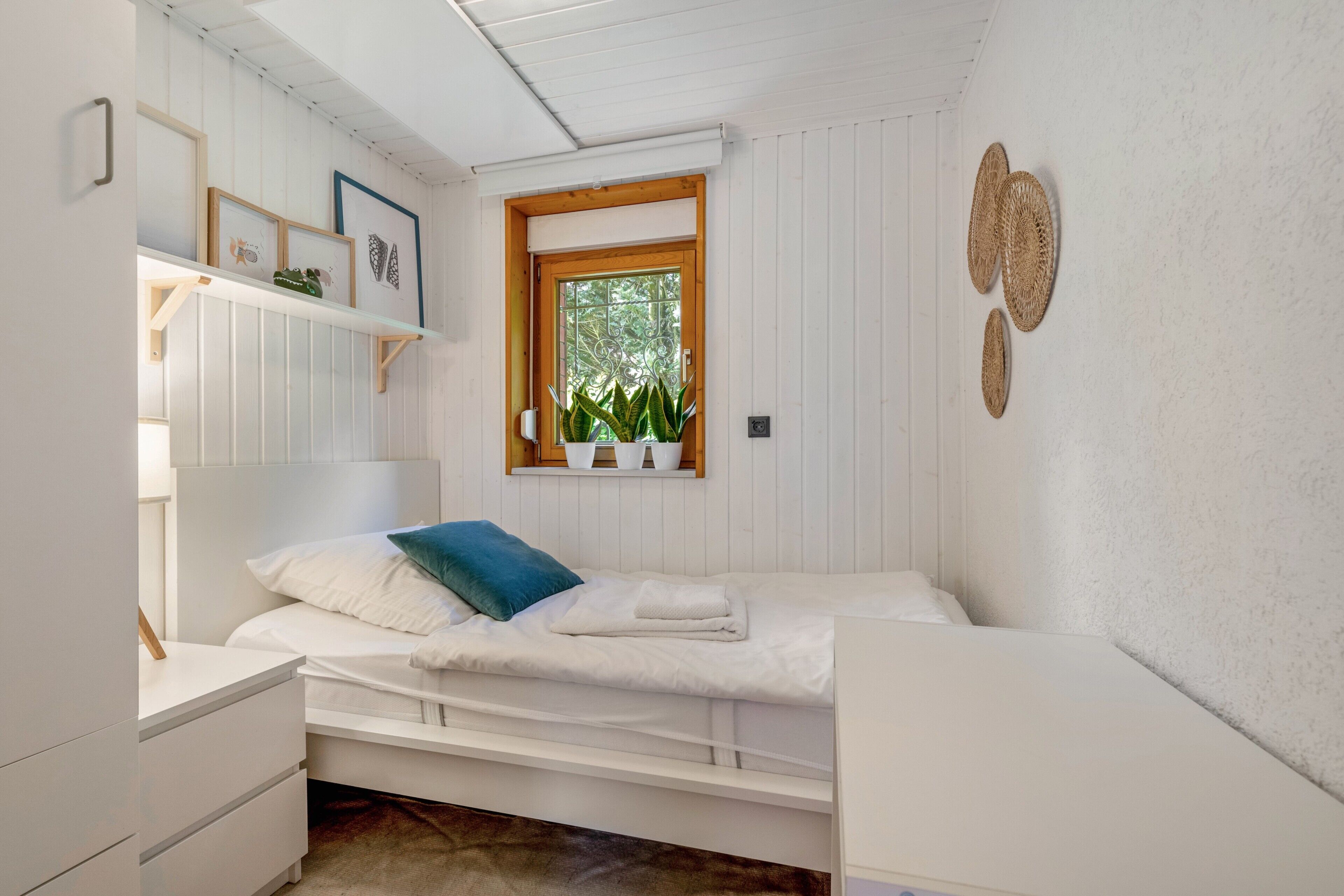 A small, white room with a single bed, a nightstand, a window with plants, and wall-mounted decor.
