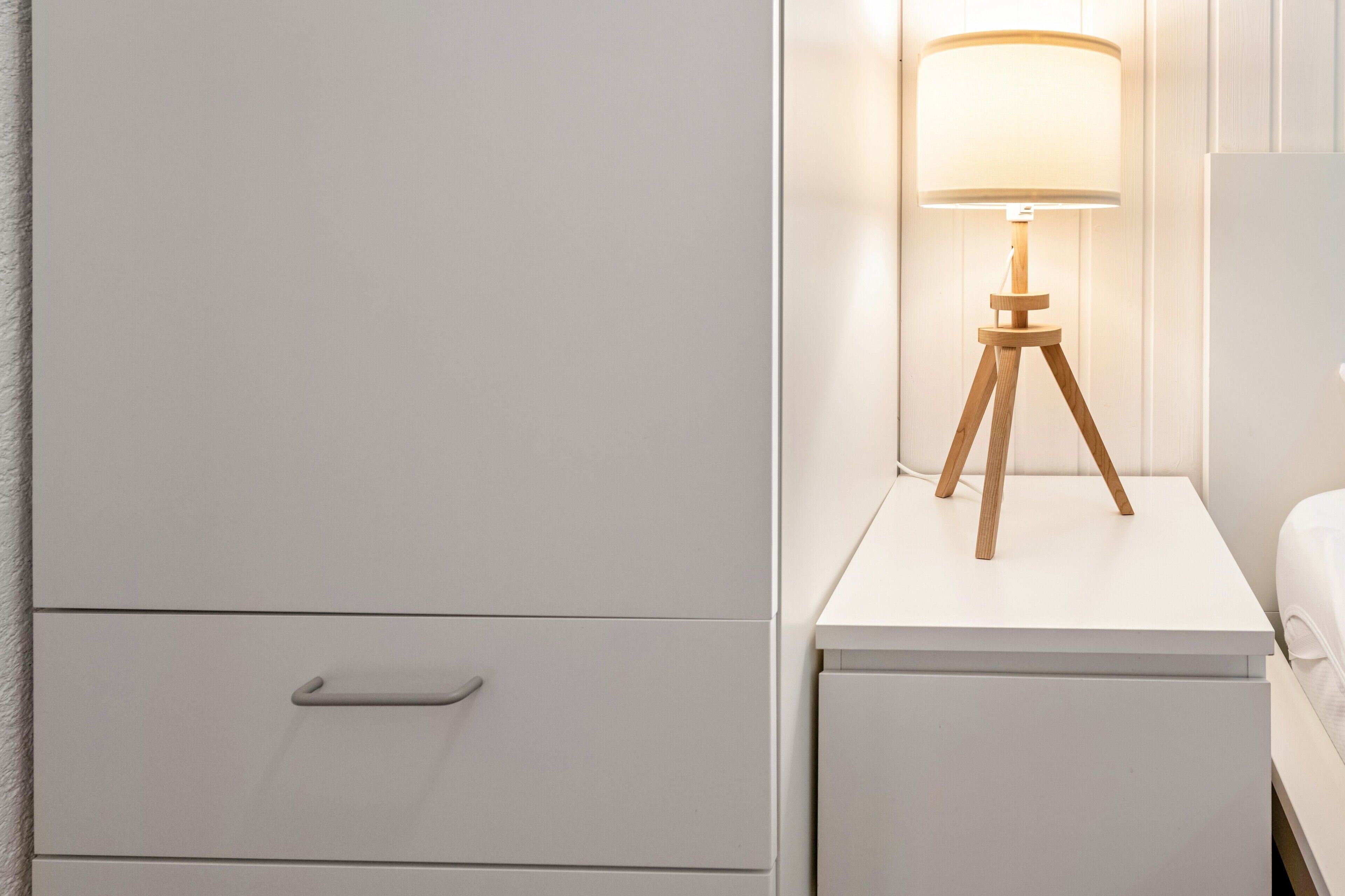 A white bedside table with a lamp, a drawer, and a bed.
