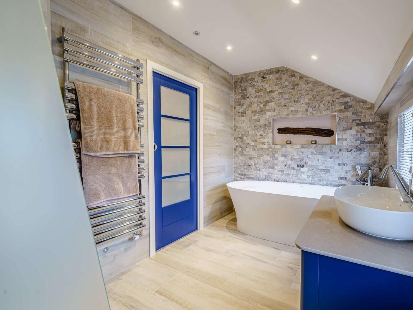 A modern bathroom with a freestanding bathtub, a towel rack with towels, a blue door, and a stone-patterned wall.