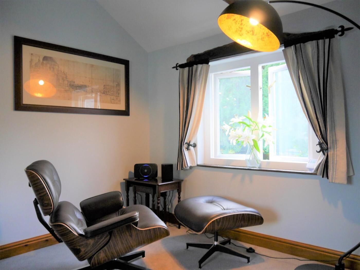 A room with a large window, a framed picture on the wall, a desk with a computer, and two chairs.