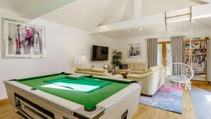 A pool table in a well-lit room with a high ceiling, a sofa, a television, and a bookshelf. - 4 Bed in Okehampton (oc-83785) (Okehampton)