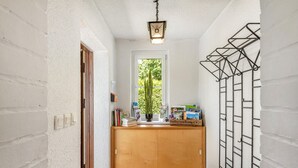 Ein schmaler Flur mit weißen Ziegelwänden, einer Holztür, einem kleinen Fenster mit Blick ins Grüne und einem Regal mit Büchern und einer Kiste.