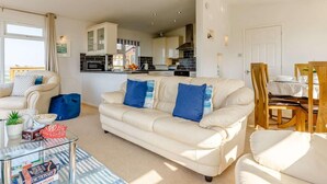 A modern living room with a sofa, a glass coffee table, and a dining area with wooden chairs. - 3 Bed in Torpoint (oc-83792) (Millbrook)