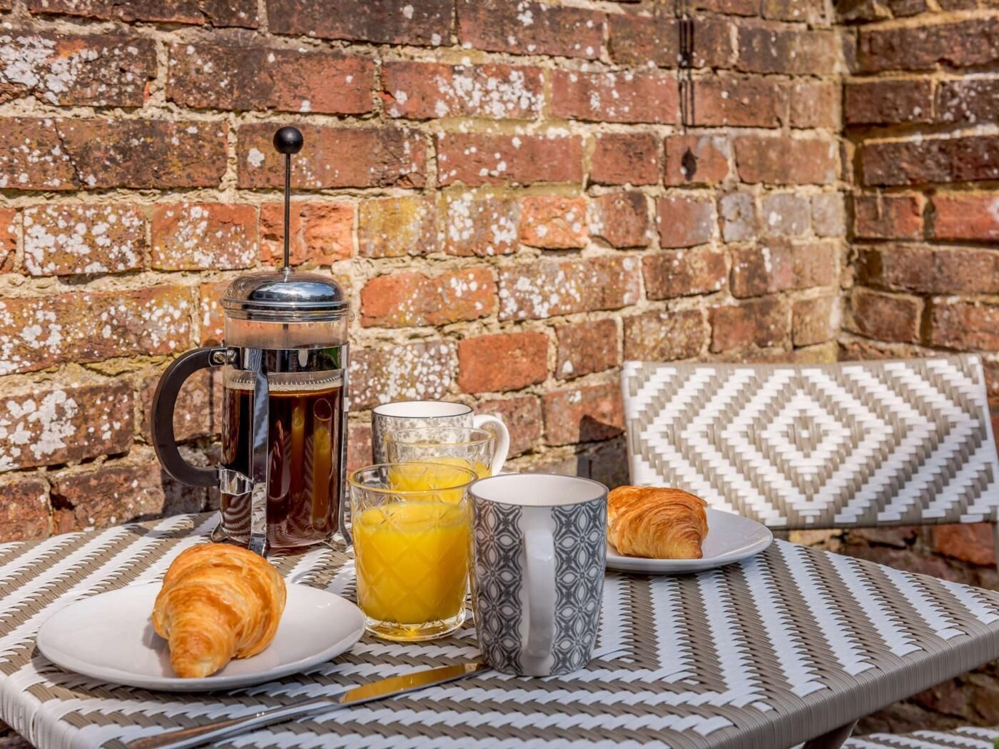 A breakfast setup with a coffee pot, a glass of orange juice, a croissant, and a mug on a woven table against a brick wall.