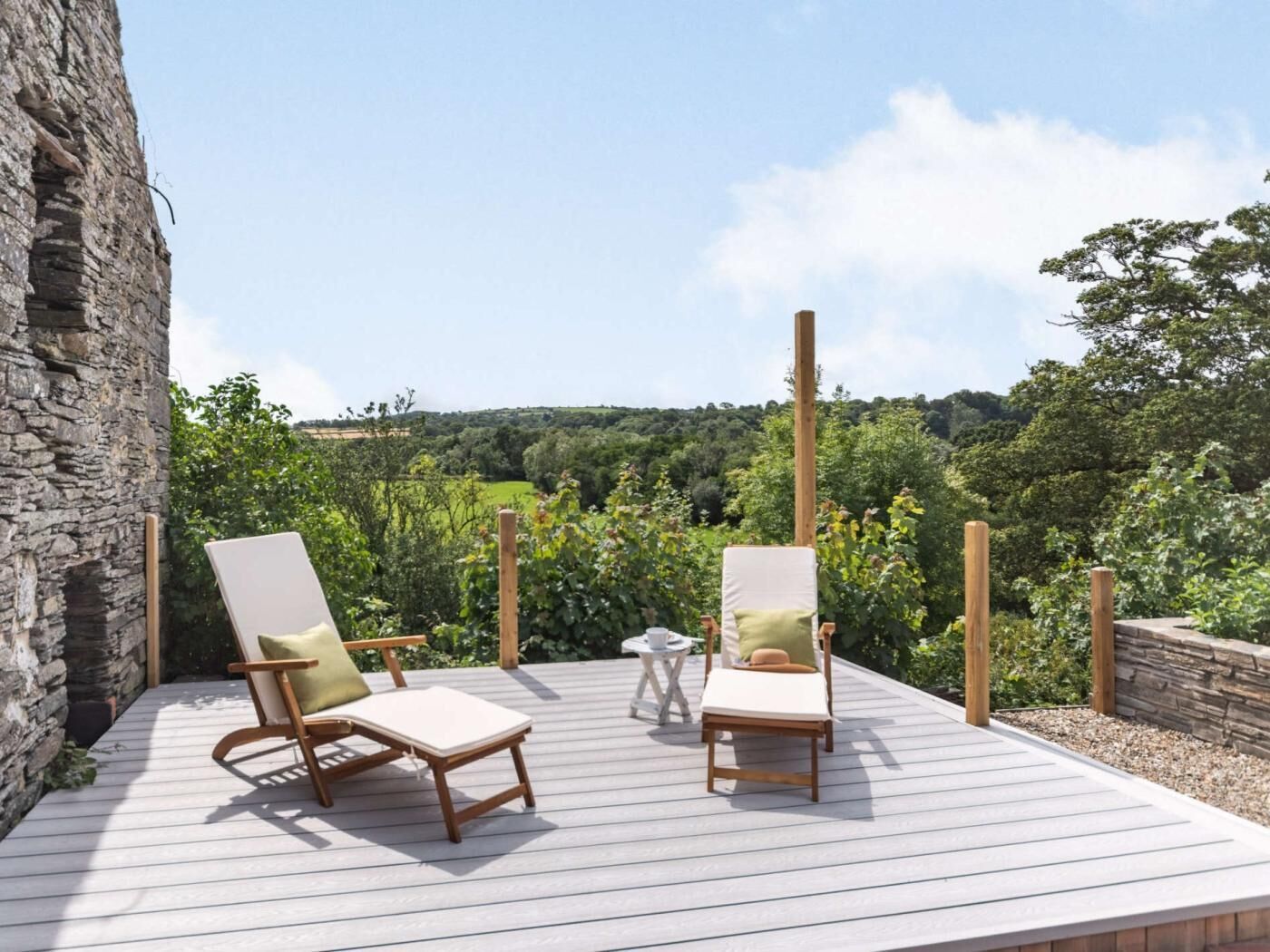 A deck with two wooden lounge chairs, a small table, and a stone wall.