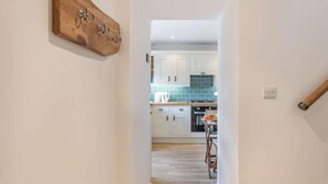 Un couloir mène à une cuisine dotée de meubles blancs, d’un pare-feu bleu et d’une décoration murale en bois.