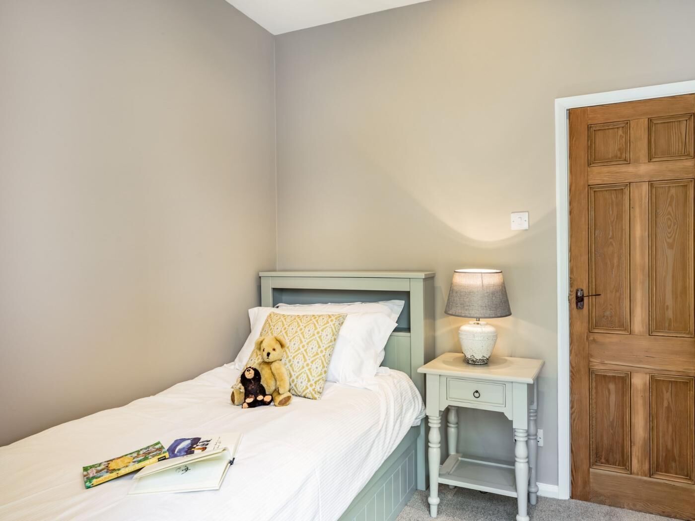 A bedroom with a bed, a nightstand with a lamp, a teddy bear, and a door.