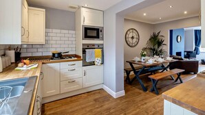 A modern kitchen with white cabinets, a microwave, and a stove. There's a dining area with a table set for two, a clock, and a potted plant. - 3 Bed in Filey (oc-84007) (Filey)