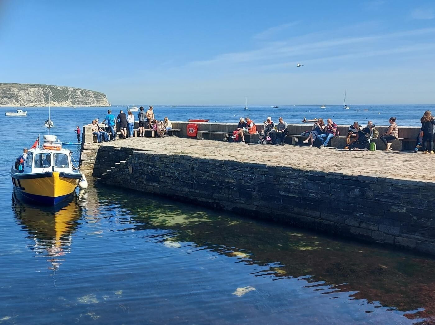 A waterfront scene with a docked boat, a stone pier, and people sitting and standing along the edge.