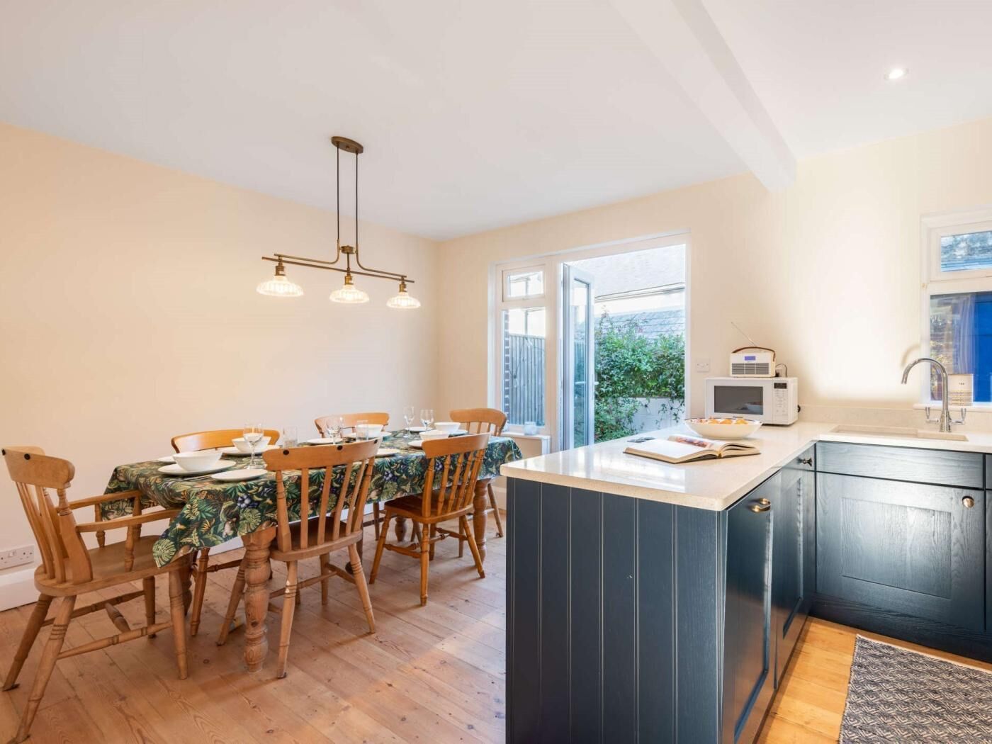 Un coin repas avec une table dressée pour quatre personnes, des chaises en bois, un luminaire suspendu et une vue sur l’extérieur grâce à une grande fenêtre.