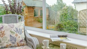 Ein gemütliches Zimmer mit einer gepolsterten Bank, einem kleinen Tisch mit Tablett und zwei Tassen sowie einem Fenster mit Blick auf einen Garten und eine Holzkonstruktion.