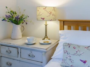 A bedroom with a wooden headboard, a bedside table with a lamp, a vase with flowers, a cup, and a plate with a biscuit. - 2 Bed in Grange-over-sands (oc-85284) (Grange-over-Sands)